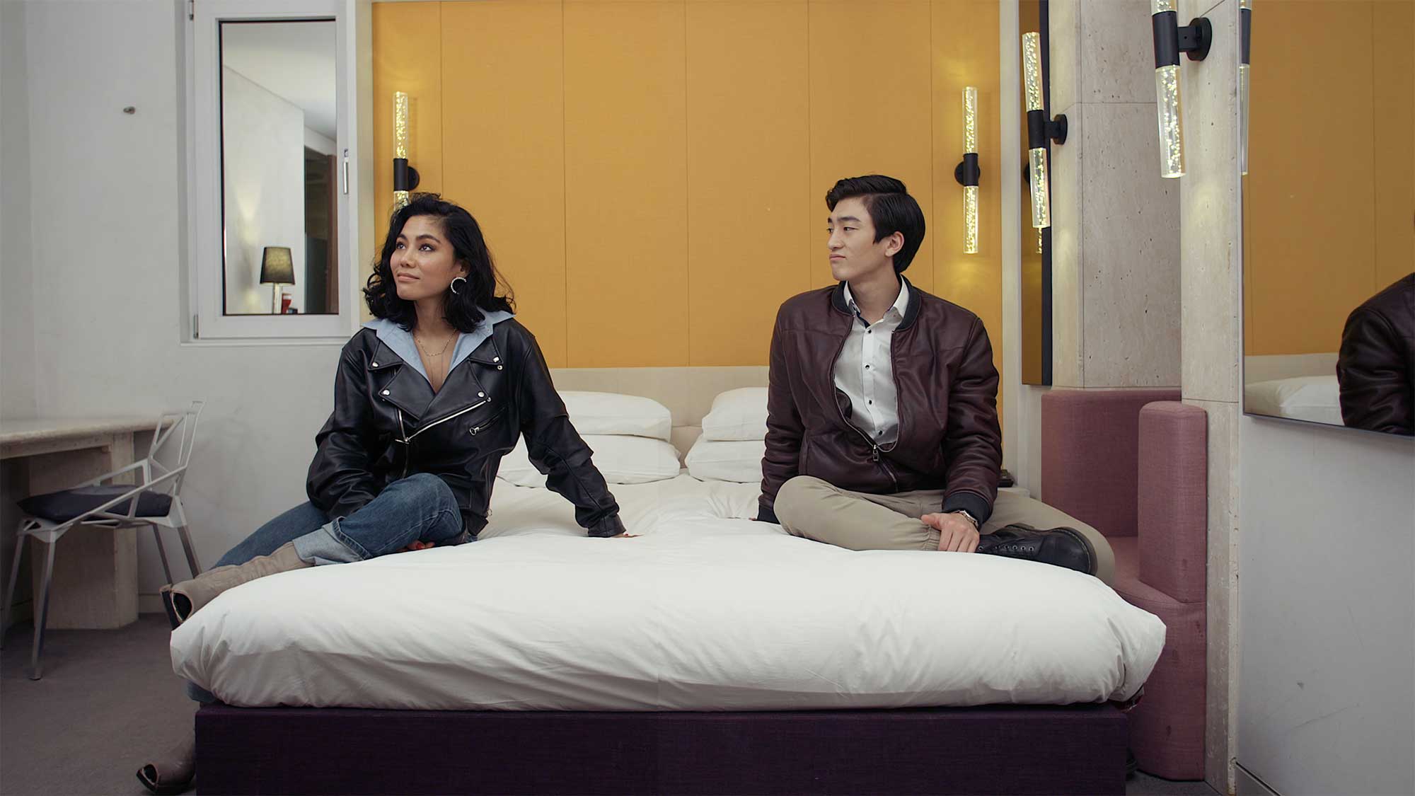 A young couple sits on opposite sides of a bed in a motel room, contemplating whether they like the place or not, in Seoul, South Korea.