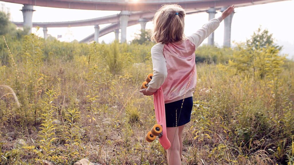 Girl with skateboard walks through grassland next to flyovers crossing Han River in Seoul, South Korea, chasing dragonflies.