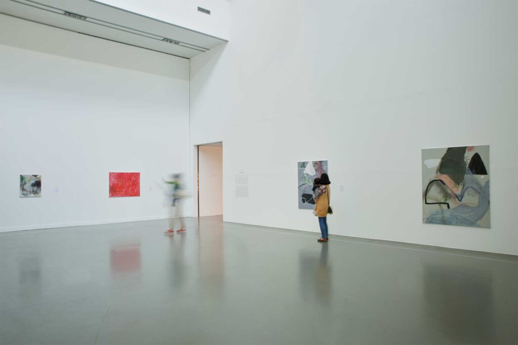 Larger-scale paintings displayed in white exhibition space with visitors passing by, part of Irony & Idealism exhibition at Gyeonggi Museum of Modern Art, South Korea.