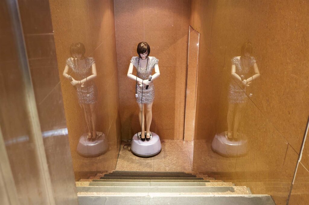 A human-like plastic doll is placed at the bottom of a bar staircase in Seoul, South Korea, dressed in shiny, sparkly attire, holding a mic with a power cable draped around her neck.