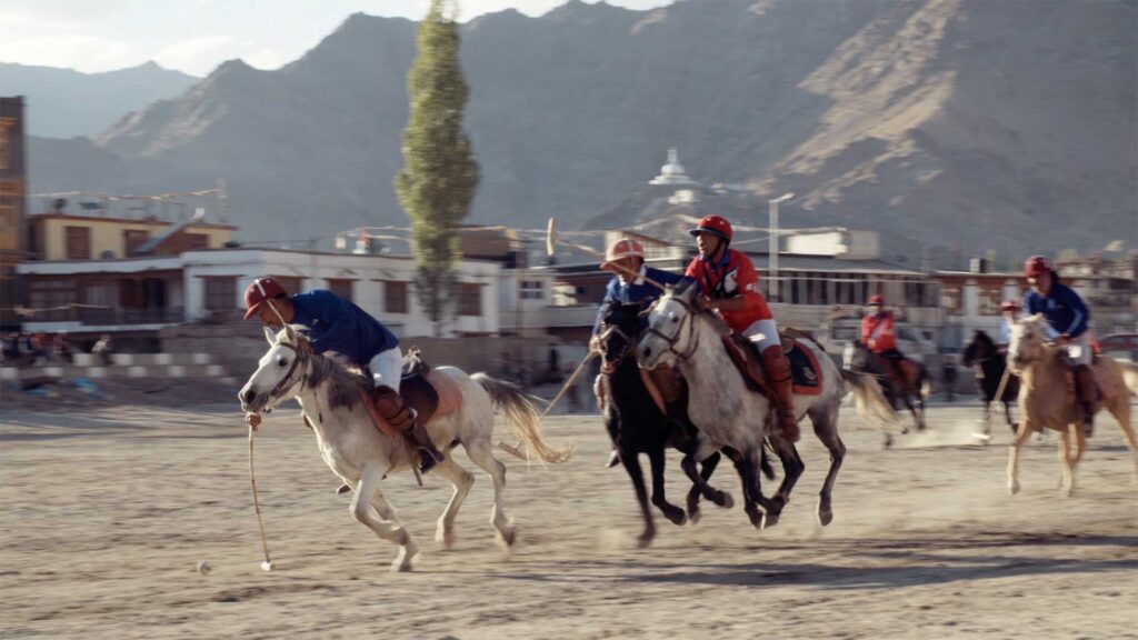 A polo game unfolds in a large open square in Leh, Ladakh, India, with majestic mountains surrounding the city in the background.