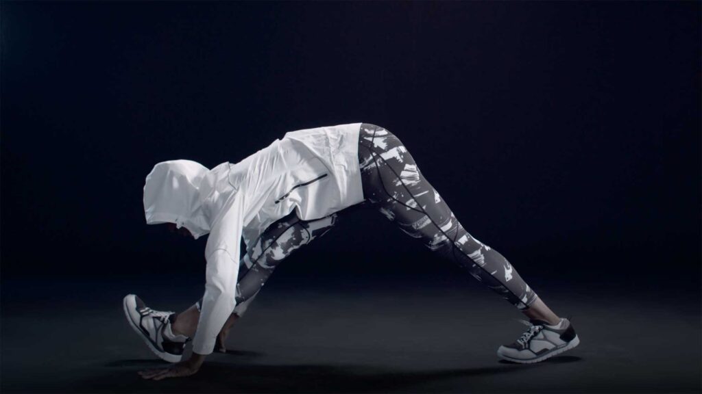 A woman of color, dressed in running shoes, patterned leggings, and a white sports jacket, deeply stretches as she prepares for a workout for a Millet sports fashion campaign.