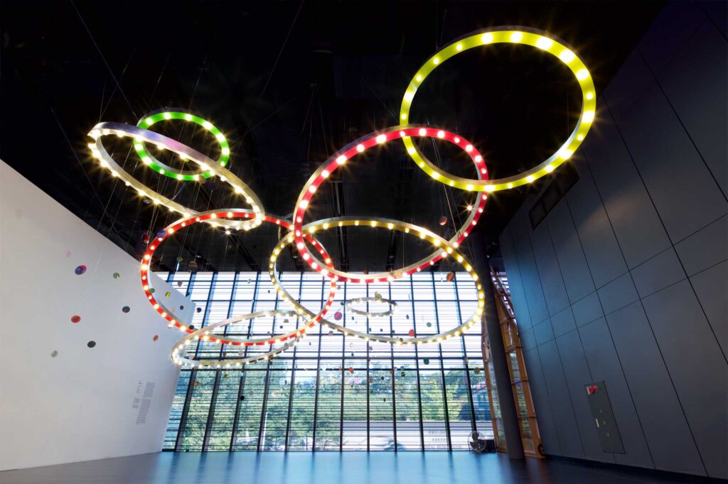 Mioon installation with circles of color and light in varying sizes hang from dark ceiling, displayed against wide open window at SUPER-SPREADER MEDIA VIRUS exhibition, Nam June Paik Art Center, Seoul, South Korea.