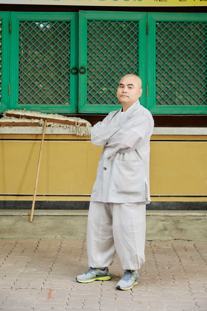 Portrait of a Korean monk in traditional attire paired with Nike sneakers, standing outside a temple against a vibrant green and yellow background in Seoul, South Korea.