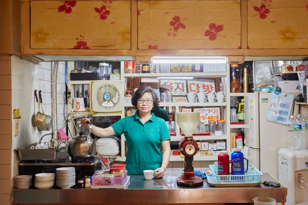 Portrait of the female owner, mid-action as she prepares to pour coffee behind the counter of a traditional Seoul coffee shop, featured in AS IT IS magazine’s issue on the beauty of aging.