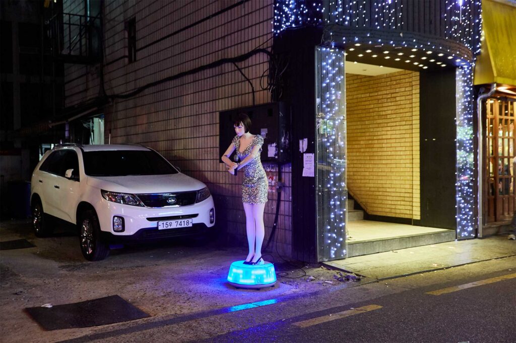 A human-like plastic doll stands outside a bar next to a parking lot in Seoul, South Korea, on a neon lid foundation, dressed in shiny attire and holding a mic.