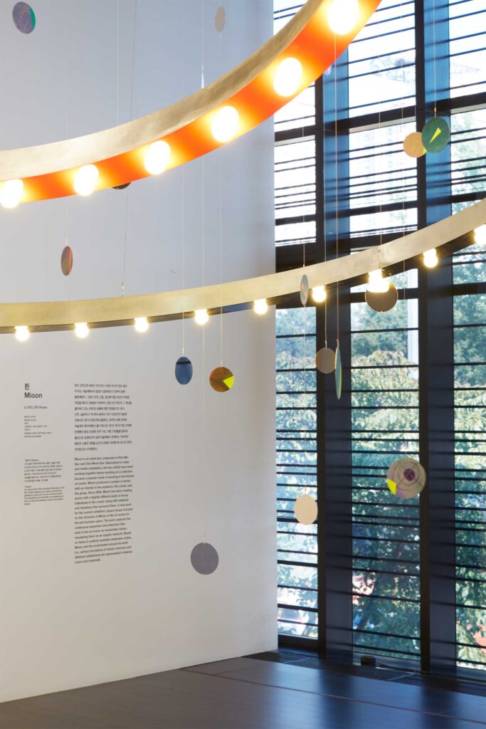 Detail shot of Mioon installation featuring circles of color and light hanging from ceiling, displayed against wide open window at SUPER-SPREADER MEDIA VIRUS exhibition, Nam June Paik Art Center, Seoul, South Korea.