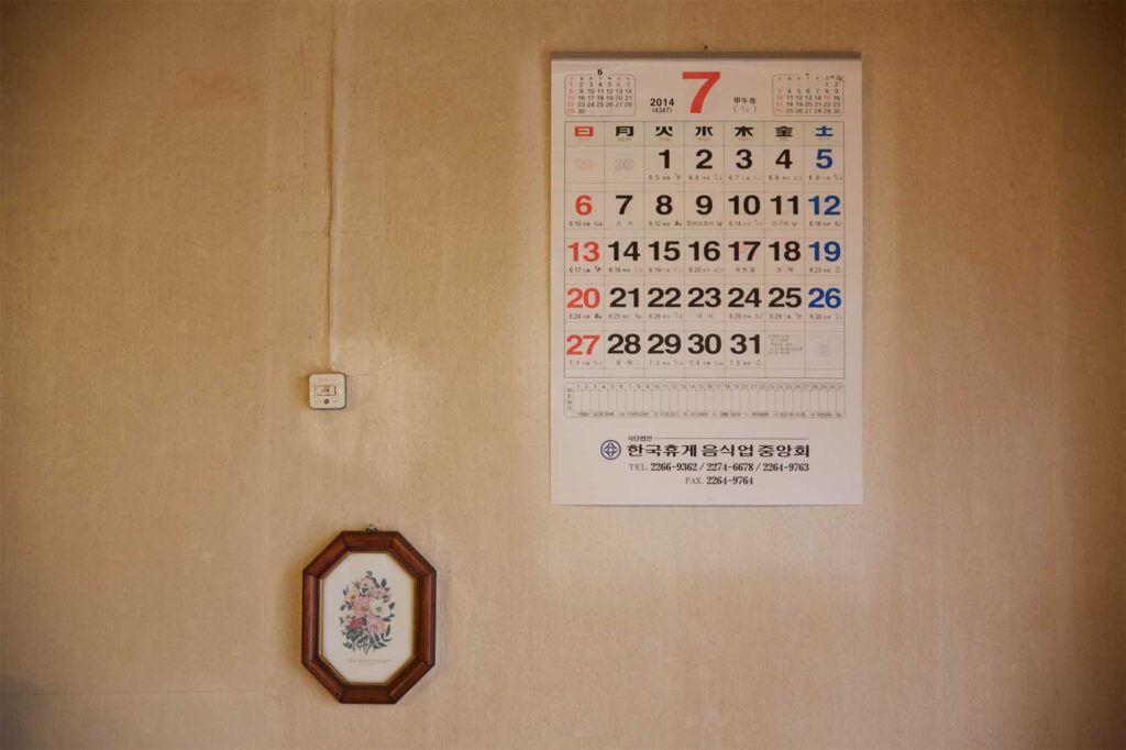 Insert shot of a yellowed wall with a calendar and framed wildflowers near a light switch in a traditional Seoul coffee shop, featured in AS IT IS magazine’s issue on the beauty of aging.