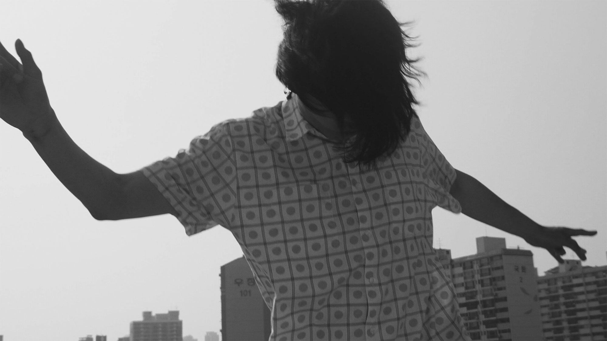 Korean musician dancing and shaking long hair while performing on rooftop of building in Seoul, South Korea, for music video "Beautiful Woman" from Tune-Up Joong-Hyun Shin: Origin tribute album.
