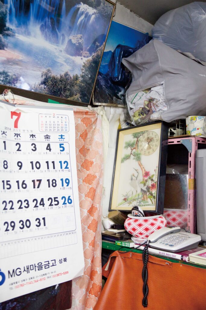 A cluttered corner with a counter holding a phone, books, and bags, surrounded by a waterfall image and calendar in a Seoul hairdresser shop, for AS IT IS magazine’s issue on the beauty of aging.