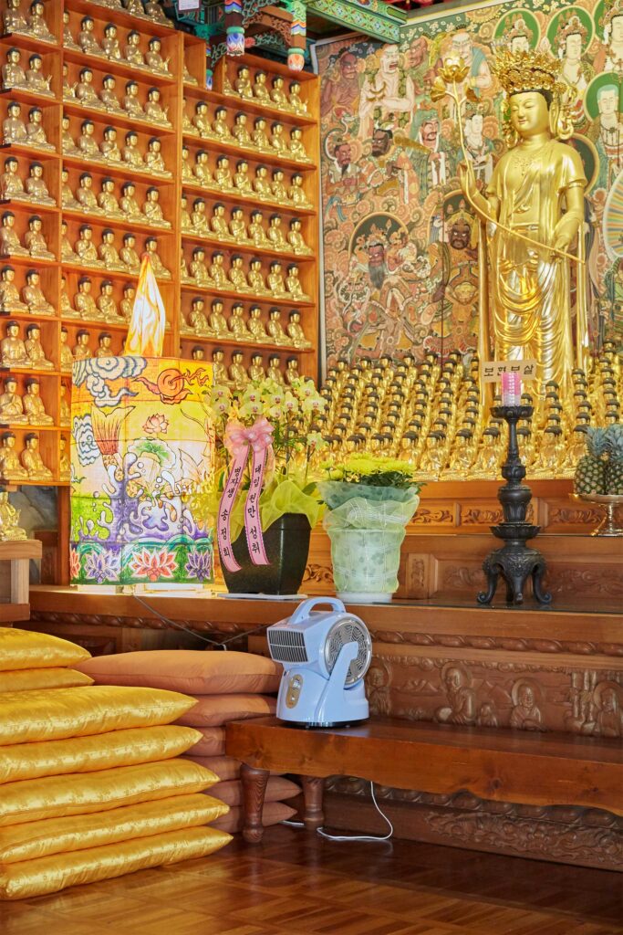 A corner of a Buddhist temple features smaller Buddha statues beside a larger one, with a blue robot-shaped ventilator adding an offbeat charm.