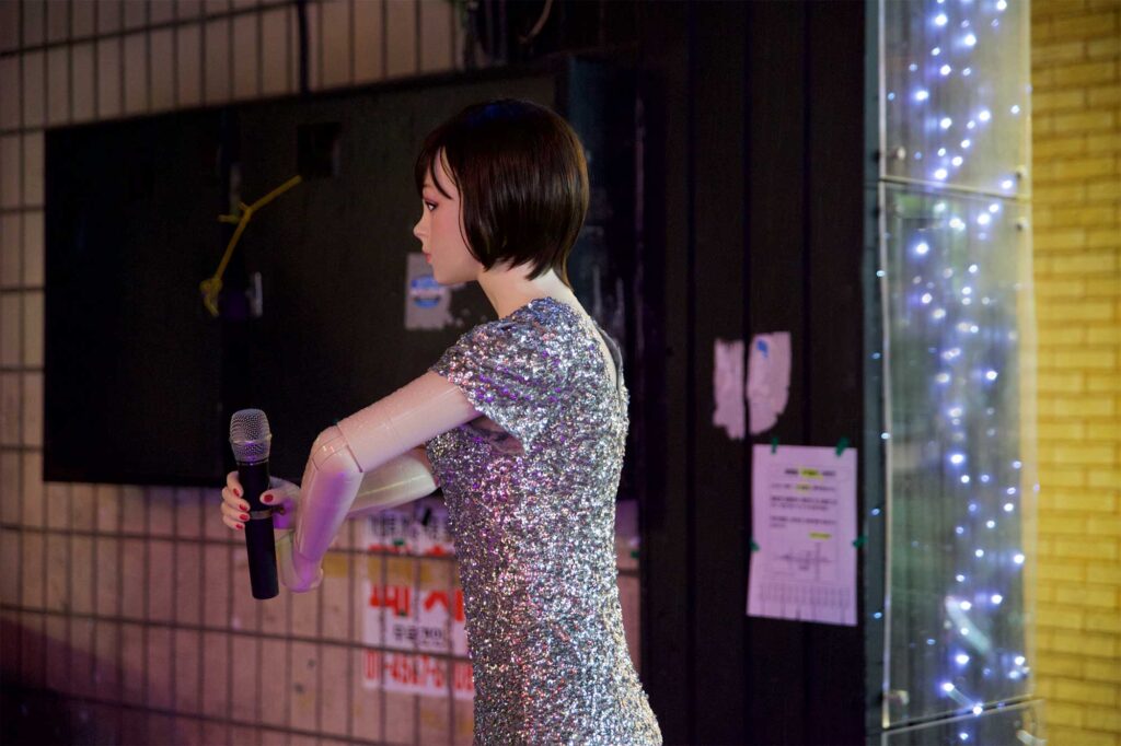 A human-like plastic doll stands outside a bar next to a parking lot in Seoul, South Korea, holding a mic, dressed in shiny attire with short sleeves revealing her plastic arms.
