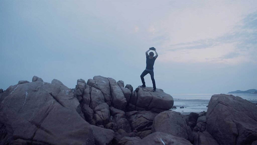A young man stands atop a coastal rock formation, smashing a stone, embodying one of the lost souls of the Sewol ferry disaster in this dream sequence for the short film Last Letters.