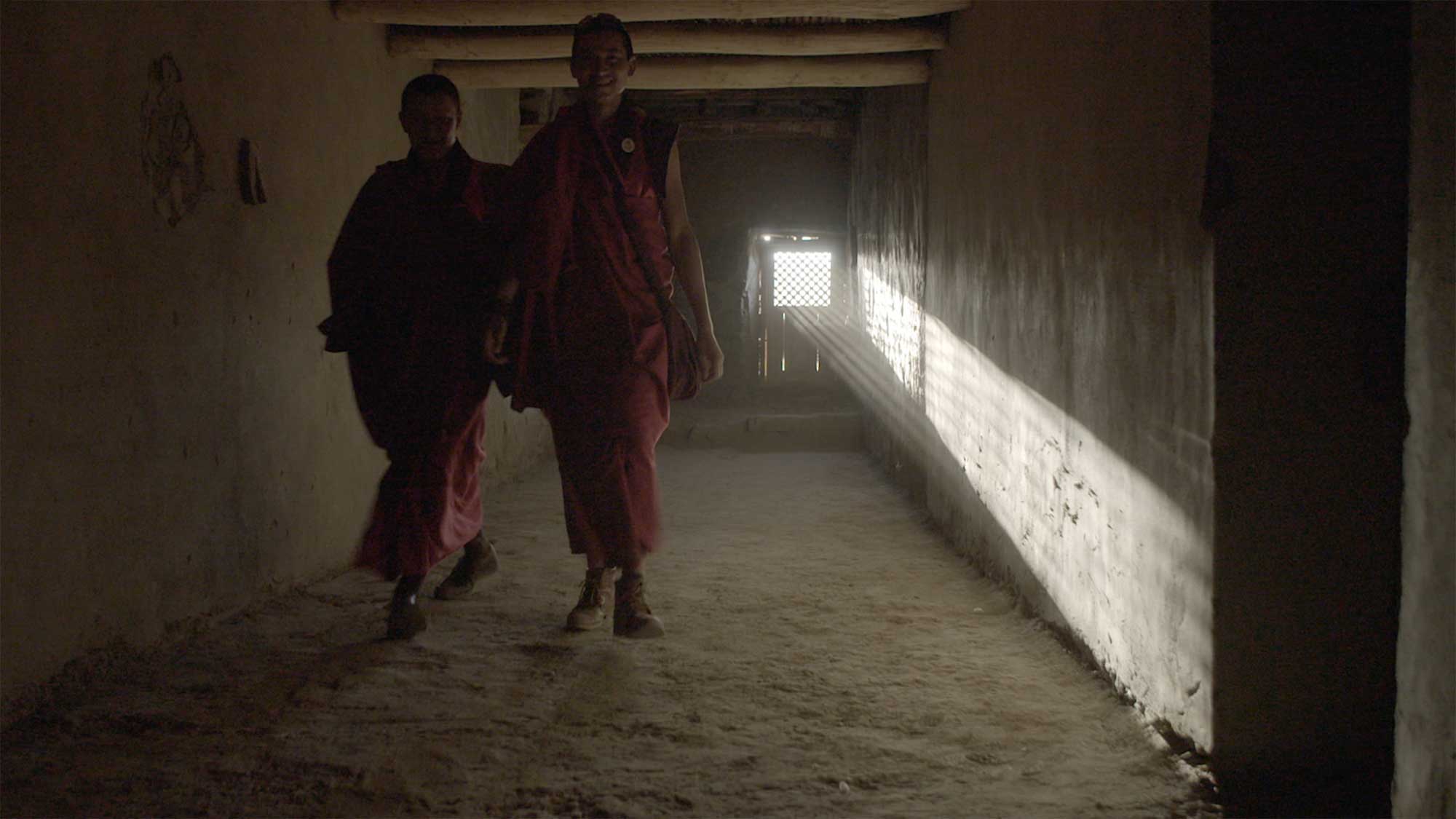 Two young monks walk through a dark temple alleyway in Leh, Ladakh, India, with sun rays streaming through a small window, casting light on the wall