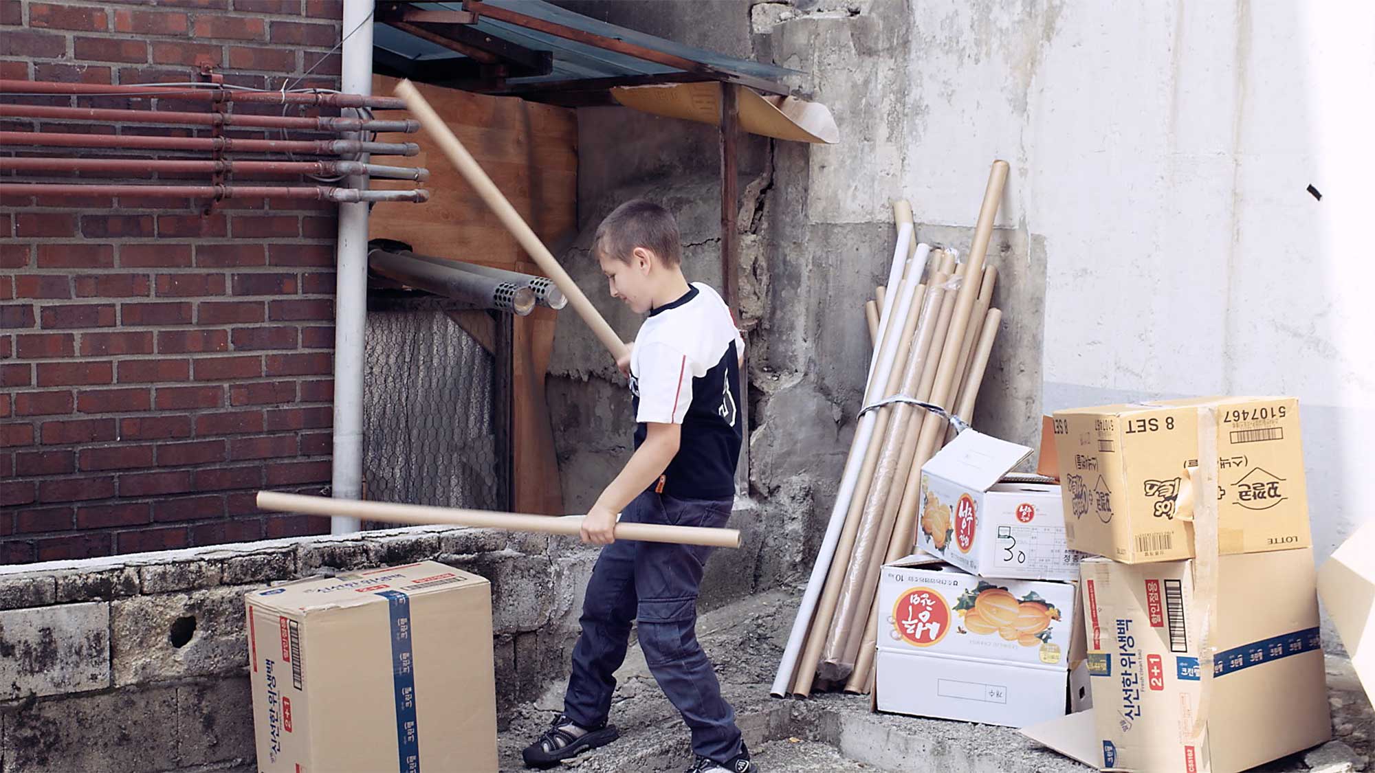 Young boy in older neighborhood of Seoul, South Korea, about to build architectural structure with cardboard, gets distracted and plays drums on boxes.