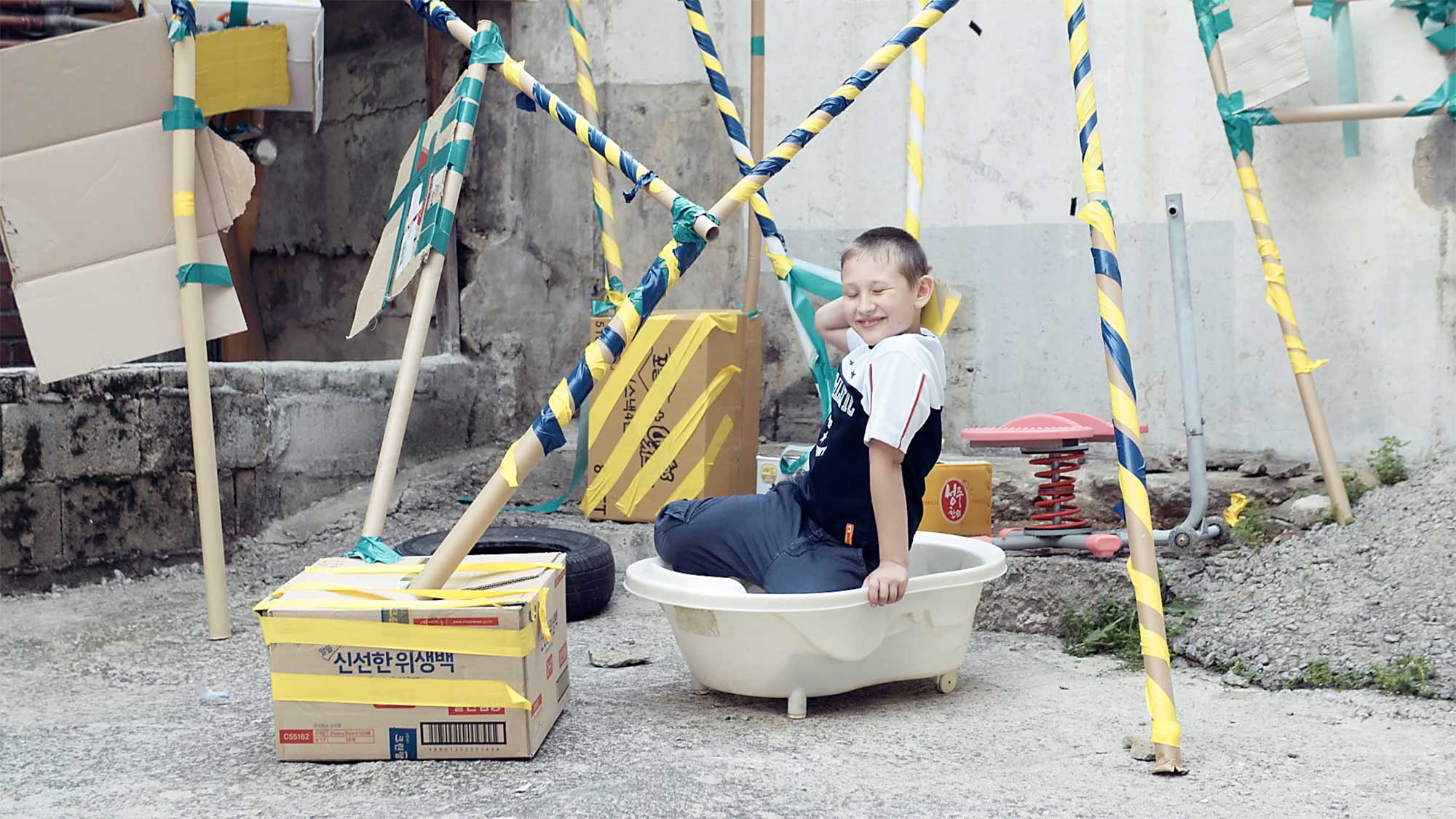 Young boy in older neighborhood of Seoul, South Korea, sits in self-built cardboard structure, pretending to take a bath as if in a real bathtub.