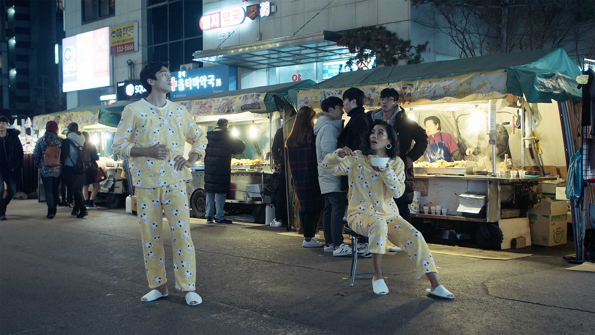 A young couple, dressed in pajamas, enjoys a late-night snack in the middle of a bustling night market in Seoul, South Korea.