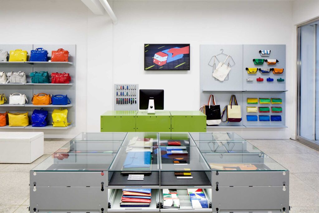 In the foreground of Freitag MMMG Seoul, South Korea, a sleek display counter showcases accessories, while the background features a cashier counter flanked by wall shelves displaying bags and accessories.