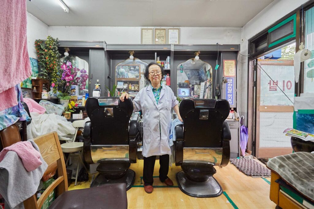 Portrait of an older female owner standing between two barber chairs in her cluttered Seoul hairdressing salon, featured in AS IT IS magazine's issue on the beauty of aging.