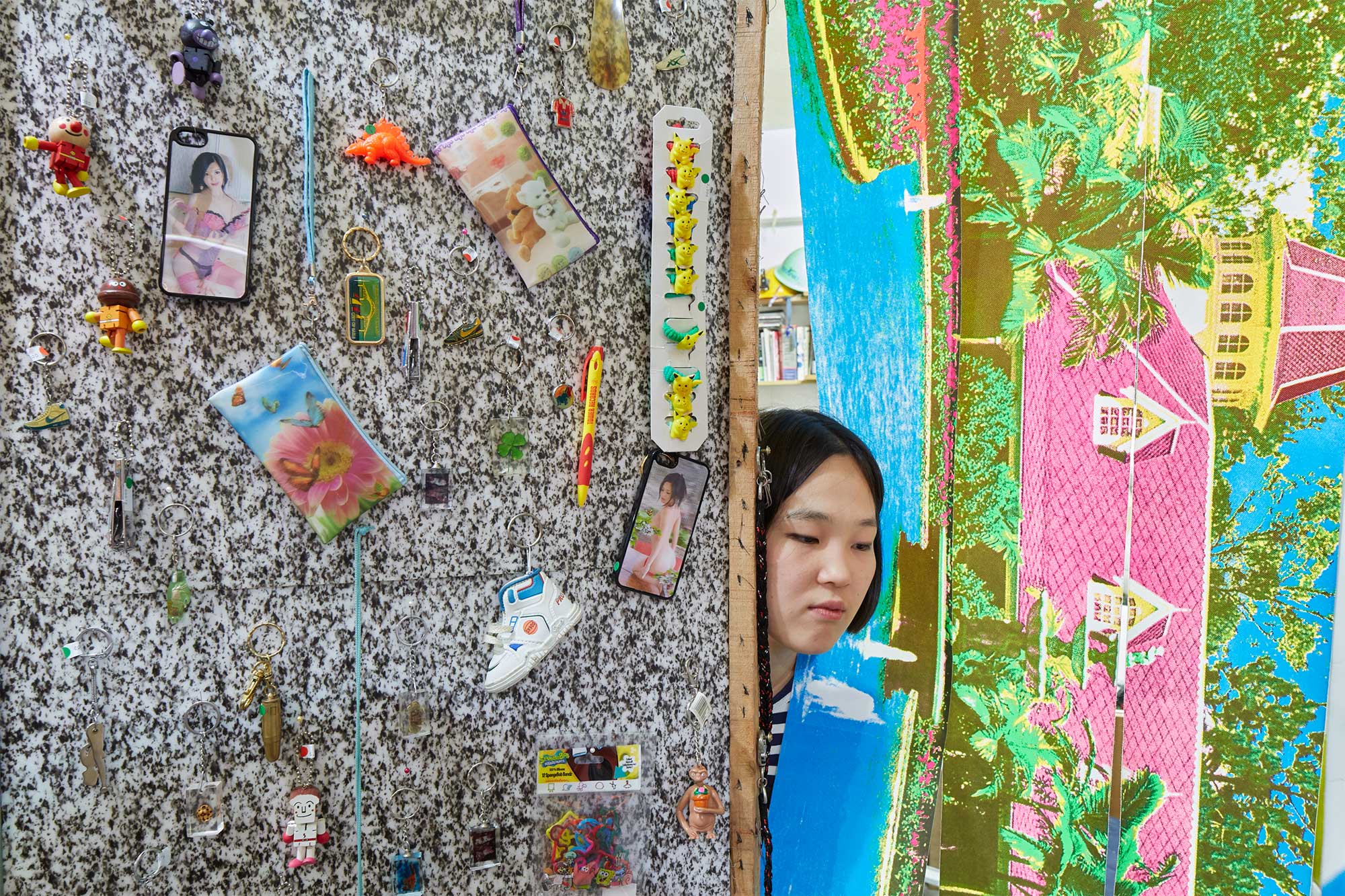 The store owner of a small kinky accessory shop in Seoul, South Korea, peeks through a colorful curtain featuring a posh offbeat house and garden landscape.