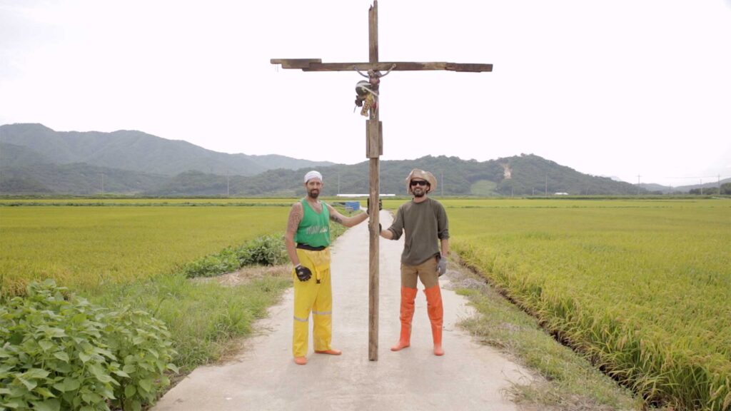 Two artists from residency in small village Yangjiri near Cherwon, Kangwando Province, holding up cross with Jesus figure nailed to it on road between rice fields in short film ART ATTACK.