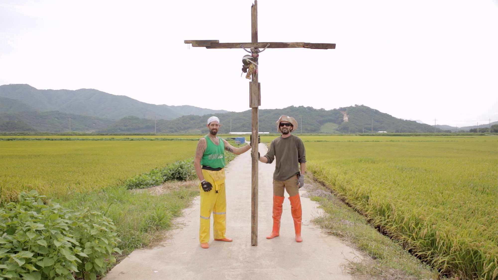 Two artists from residency in small village Yangjiri near Cherwon, Kangwando Province, holding up cross with Jesus figure nailed to it on road between rice fields in short film ART ATTACK.