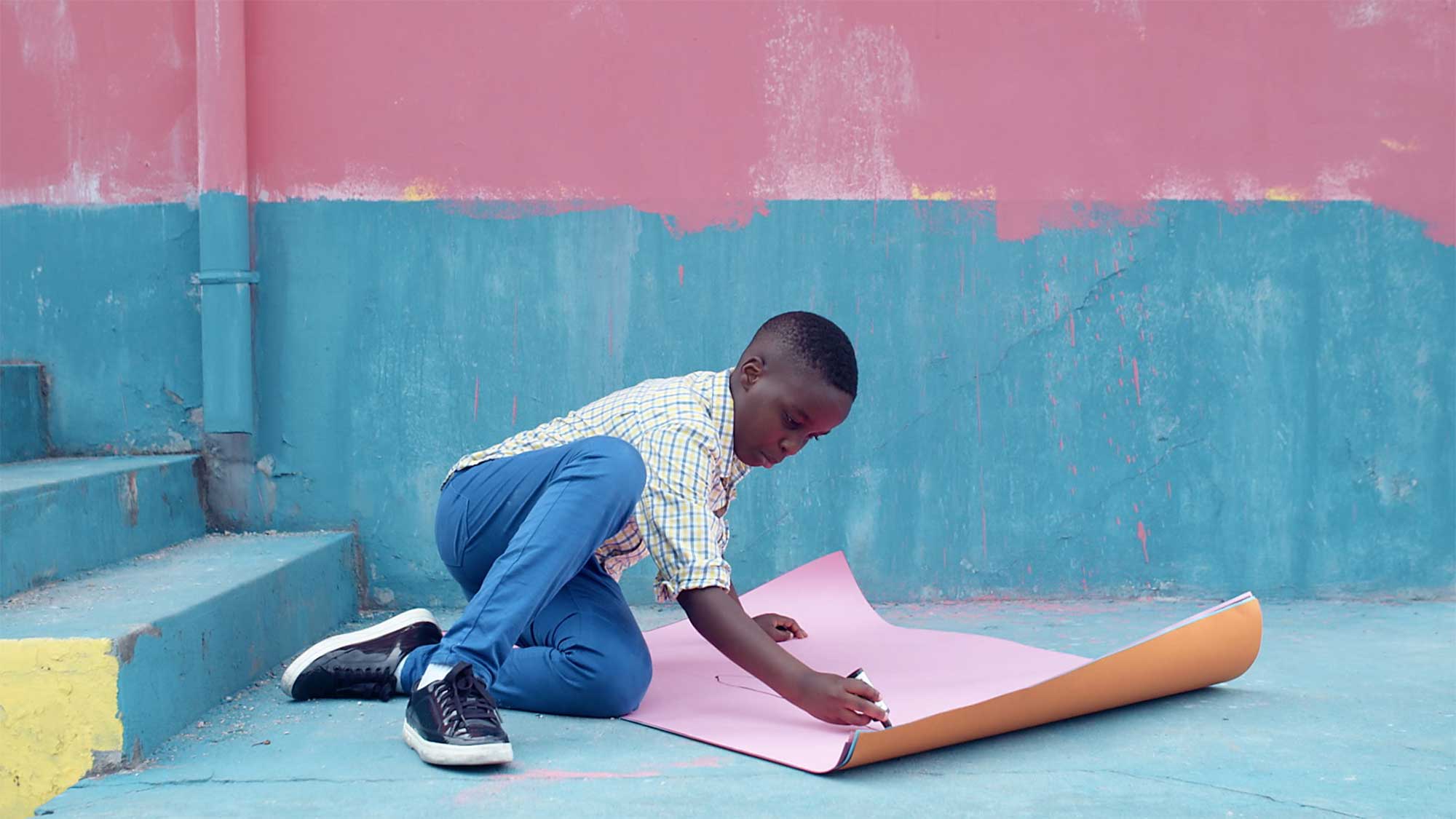 Young boy sits on colorful exterior staircase of building in Seoul, South Korea, drawing with sign pen on big cardboard paper.
