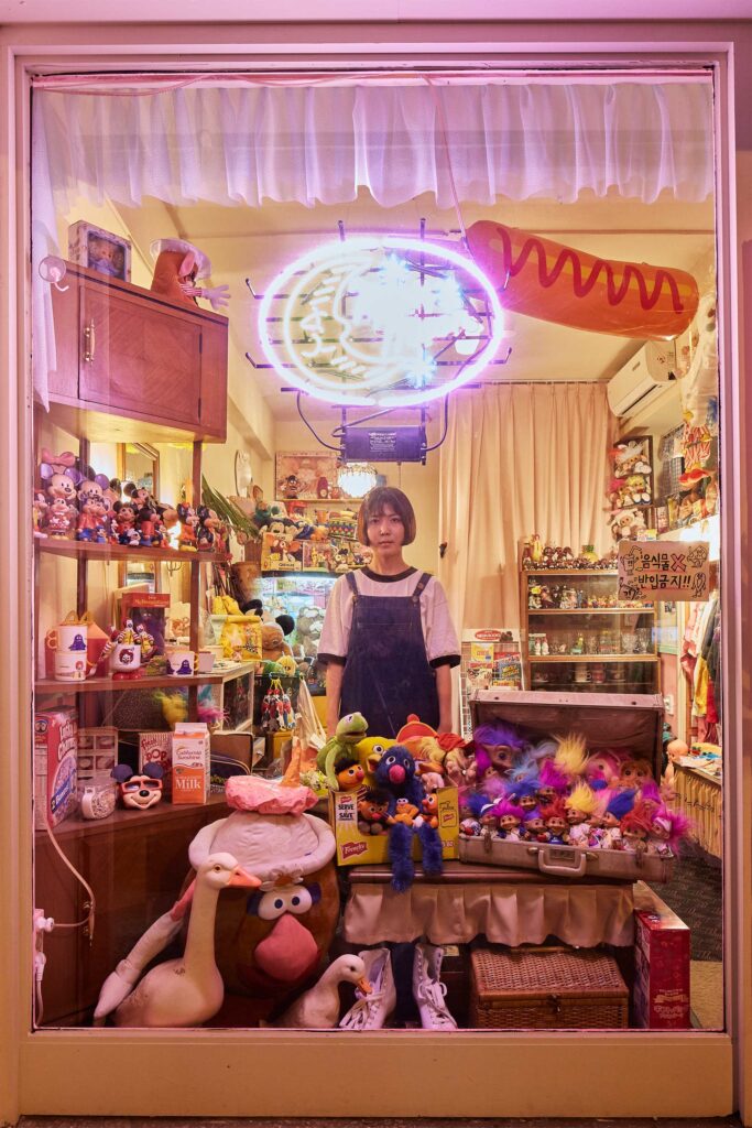 The store owner framed from outside the window of her small shop filled with whimsical puppets and comic characters in Seoul, South Korea, surrounded by colorful displays.