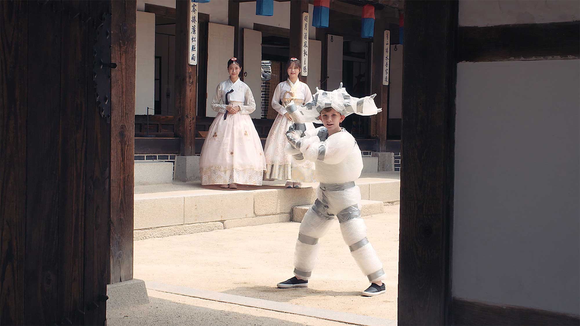 Young boy fully wrapped in bubble wrap, resembling a warrior with a bubble wrap toy gun, jumps into doorway of older temple structure in Seoul, South Korea, as two girls in Hanboks stand in background.