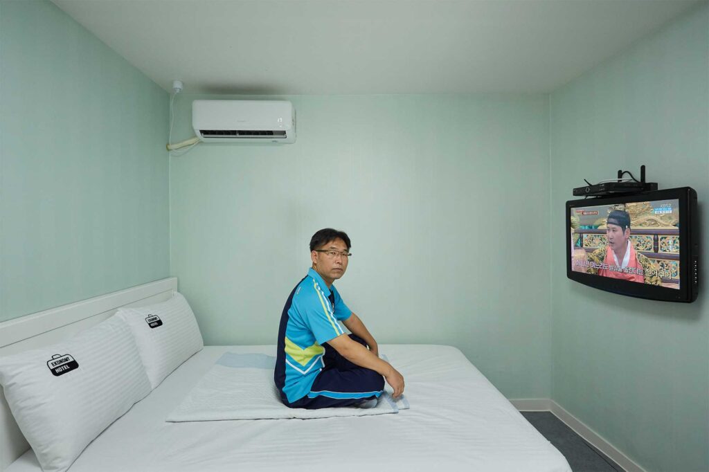 A middle-aged Korean man sits on a king-sized bed in a modest motel room with mint wallpaper, featuring only a TV and air-conditioning on the walls.