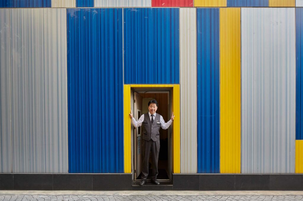 A male hotel staff stands in the side door of a boutique hotel in Itaewon, Seoul, South Korea, notable for its vibrant, striped exterior facade.