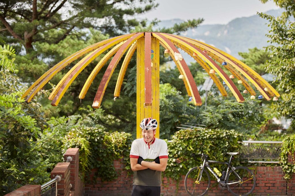 Portrait of So Wonyoung, a road cycling enthusiast, laughing joyfully beneath an umbrella frame structure along a scenic route in the Bukhansan area, South Korea.