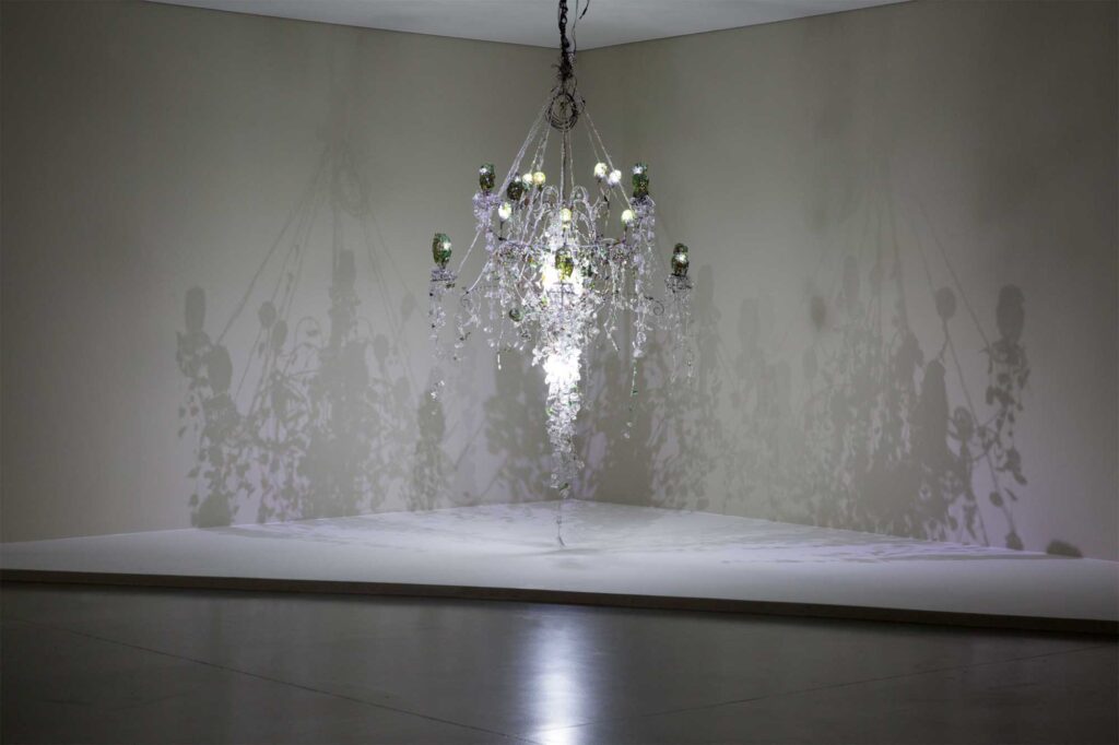 Installation art resembling chandelier overloaded with clutter at Irony & Idealism exhibition at Gyeonggi Museum of Modern Art, South Korea, casting wide shadows on wall behind.