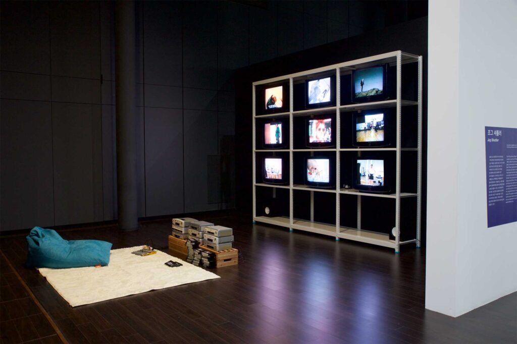 JOG SHUTTLER installation with multiple TV screens on shelf, carpet, beanbag, video cassettes, and tape players on ground in front, part of BLAST THEORY - YOU START IT exhibition, Nam June Paik Art Center, Seoul, South Korea.