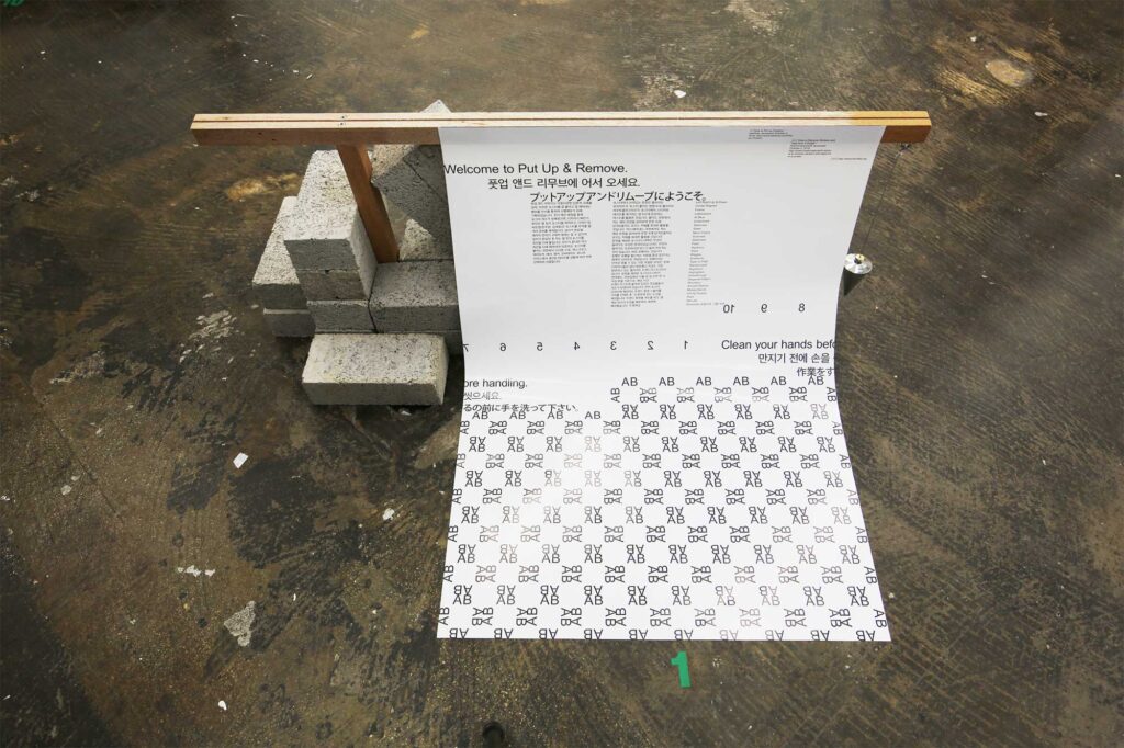 A poster half hanging on the floor, half hanging on a wooden construct stabilized by bricks for the poster exhibition in Seoul, South Korea, by Graphic Magazine as part of Propaganda Press.