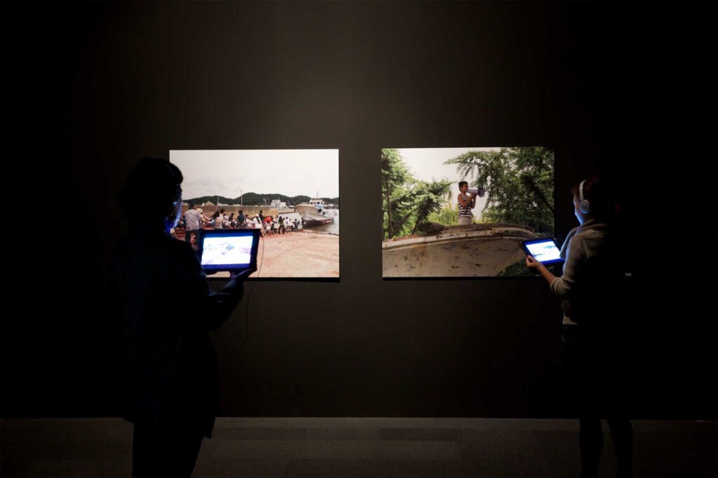 Enclosed dark room highlighting larger photo prints on wall, where visitors use iPads as audio guides to interact with images during BLAST THEORY - YOU START IT exhibition, Nam June Paik Art Center, Seoul, South Korea.