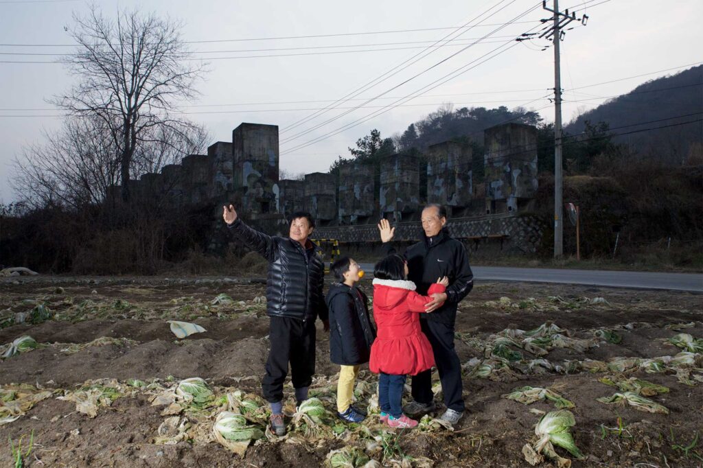 A Korean grandfather, his son, and two grandchildren pose in the early evening in front of camouflage-patterned demolition barriers lining a rural road along the former civilian control line near the Korean Demilitarized Zone.