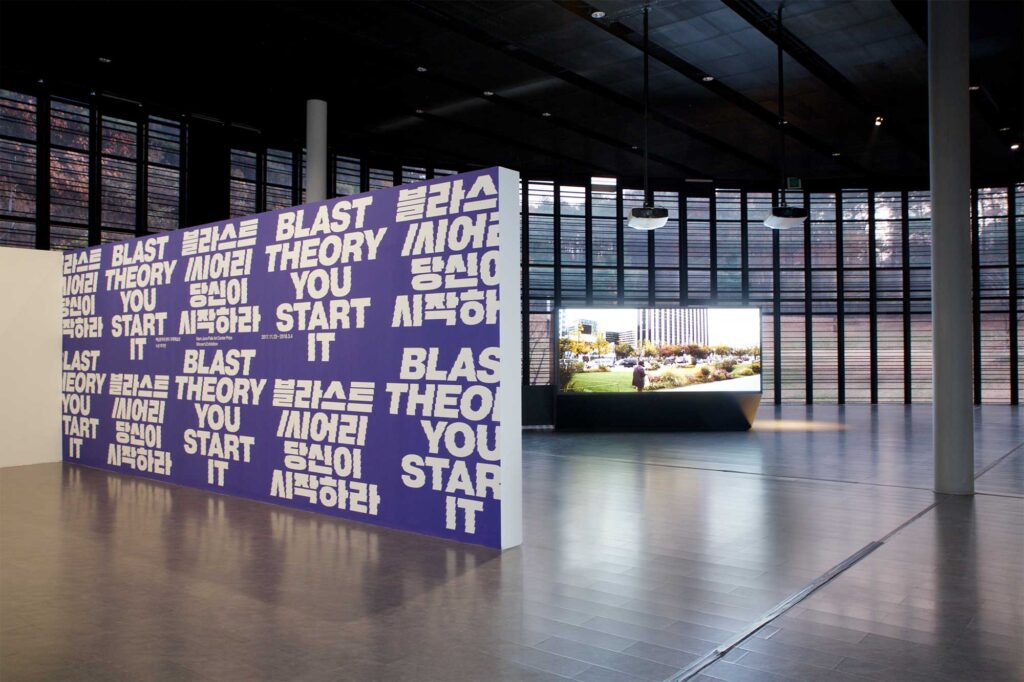 BLAST THEORY - YOU START IT exhibition at Nam June Paik Art Center, Seoul, South Korea, advertised on large dividing wall at entrance, with large screen box in spacious atrium framed by high glass facade in background.