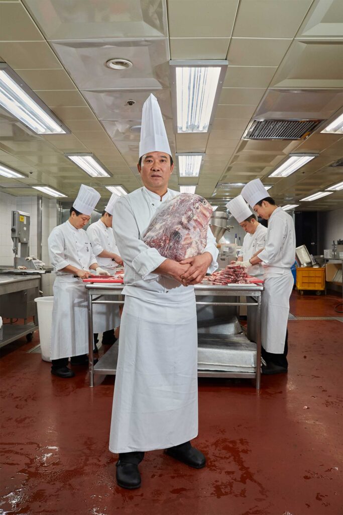 A chef from a renowned hotel chain in Seoul, South Korea, poses for a portrait holding a large chunk of meat, with kitchen staff working in the background.