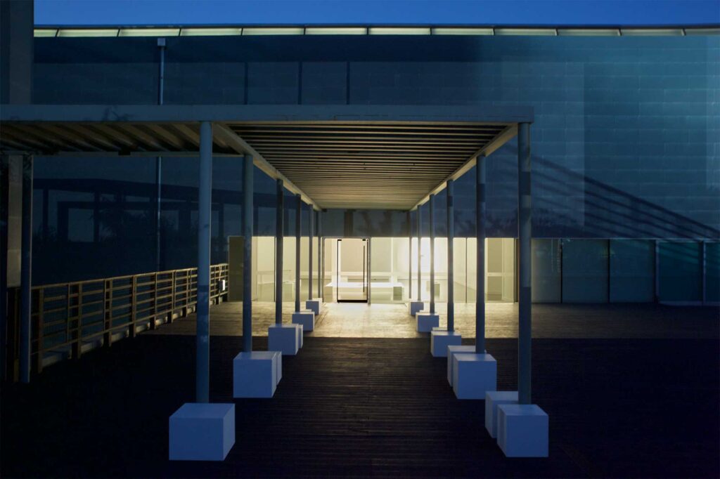 Night exterior shot of museum entrance with white architectural pavilion inside, square white chairs leading from outside to inside at Irony & Idealism exhibition, Gyeonggi Museum of Modern Art, South Korea.