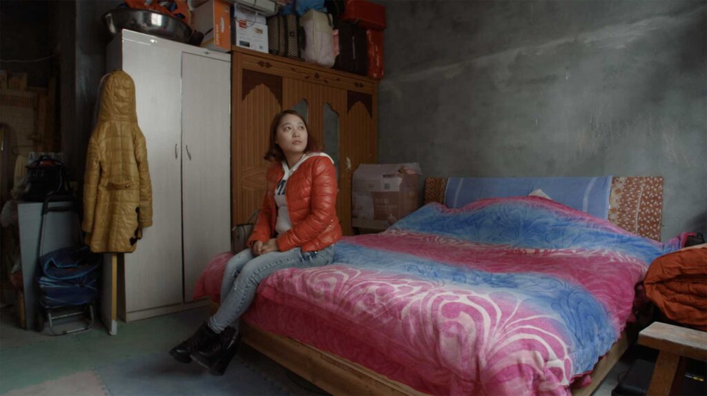 As part of a Western Union Chain of Betters campaign, a young Chinese woman sits on the edge of her king-sized bed in a cramped sleeping room with concrete walls, surrounded by furniture and personal belongings in Changle District, China.