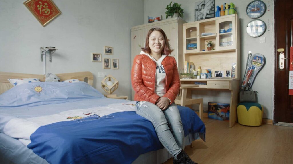 As part of a Western Union Chain of Betters campaign, a young Chinese woman sits on the edge of a bed in a newly decorated children's room in Changle District, China, featuring a desk, cupboards, a clock, and picture frames on the wall.