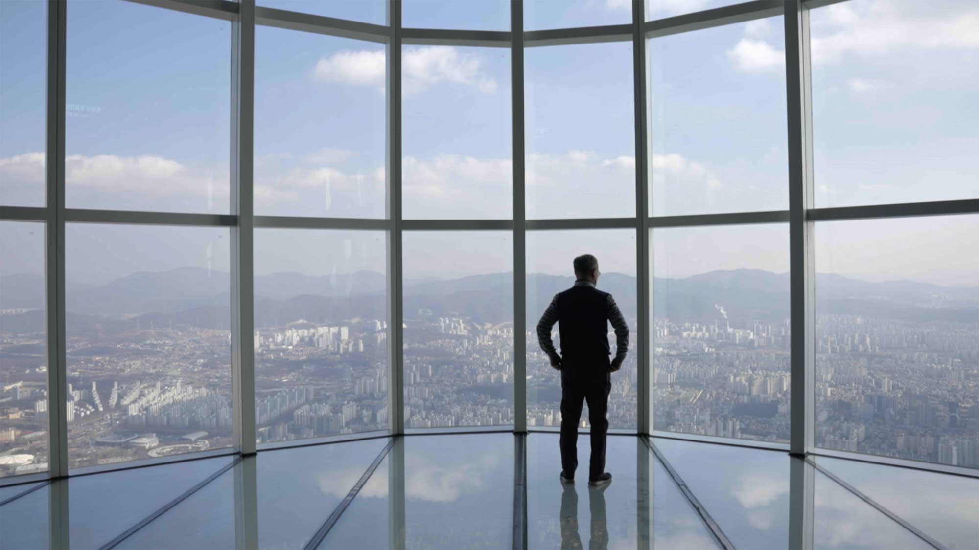 Silhouetted man stands at Lotte Tower observatory overlooking Seoul skyline under hazy sky.