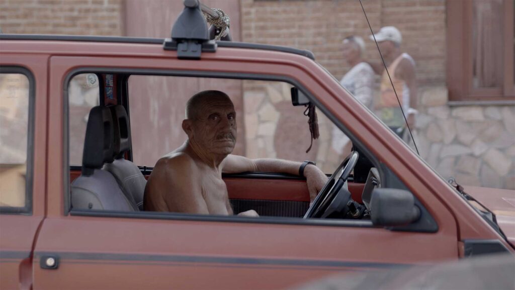 Older topless man waiting in traffic in small red Fiat, looking at camera through open window in Amantea, Italy