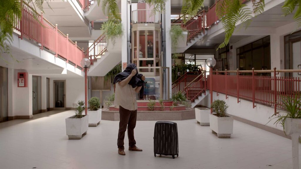 Interior filled with green plants and red-barriered balconies, man stands head covered by jacket.