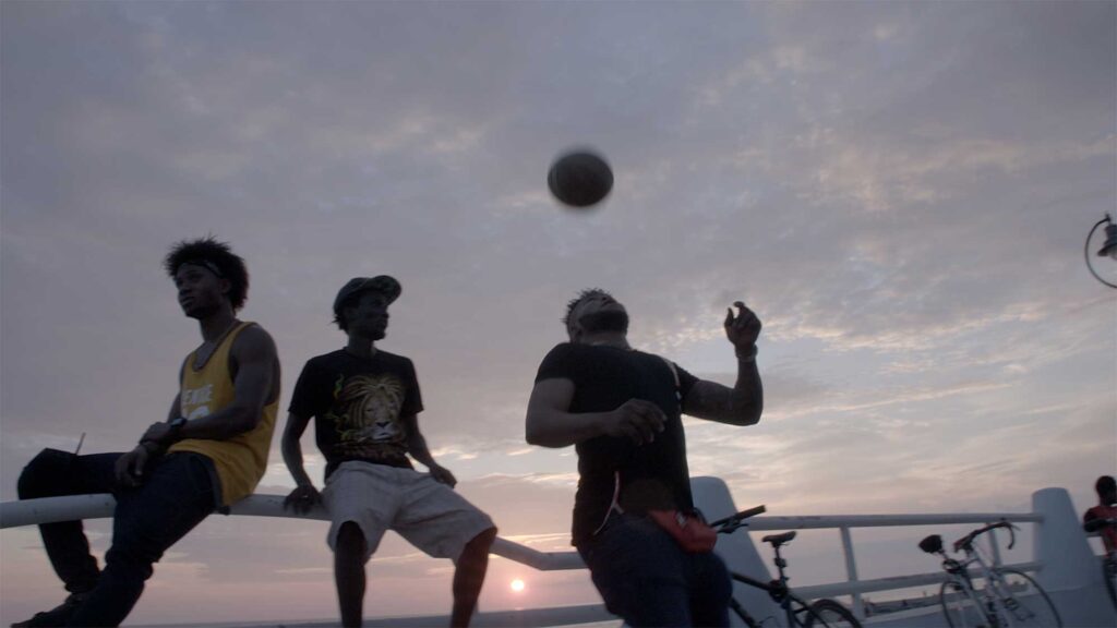 Stanley Eboigbe and two other African immigrants, Stanley dribbling ball with head up as sun sets over ocean in Amantea.