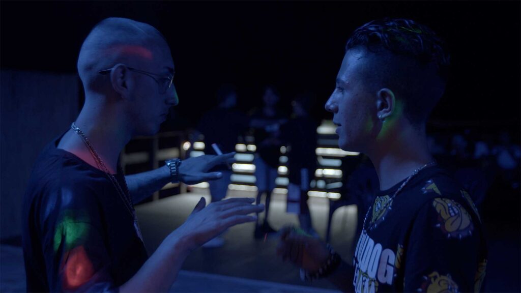 Daniele Chiodo out at night in club talking to friend, wearing gold chain and cool glasses.