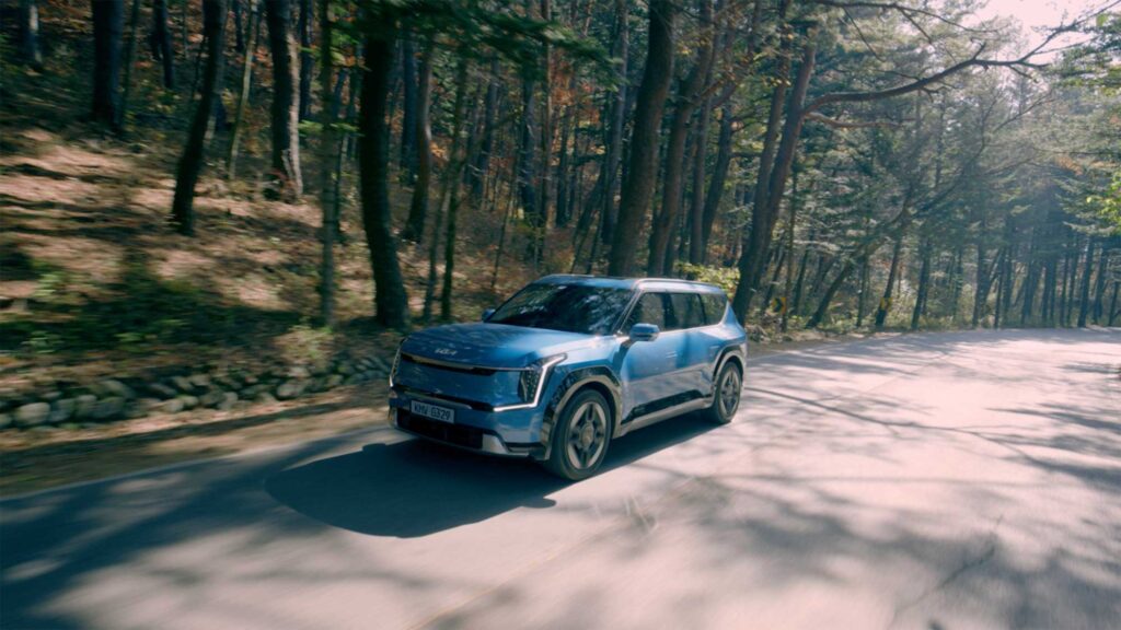 KIA EV9 driving along sunlit forest road.