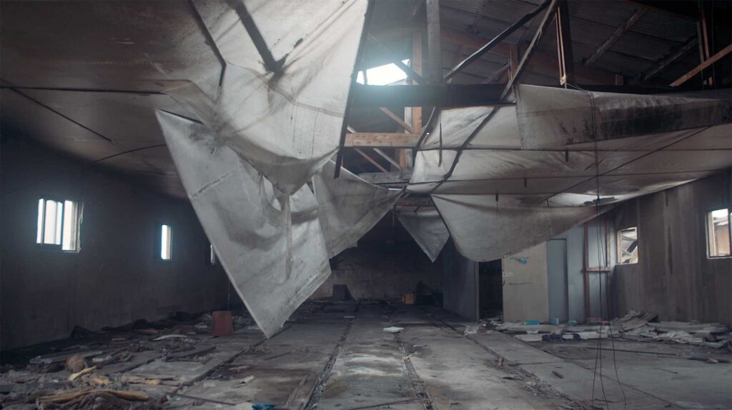 Abandoned factory hall with hanging fabric remnants and damaged roof structure reflecting collapse of industrial labor spaces in South Korea