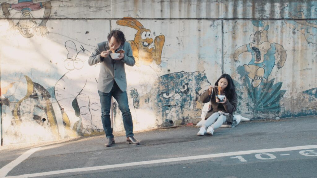 Gyungsung Lee and Korean actress sharing street meal in Seoul during Goethe Institut Seoul promo film reflecting everyday life urban culture and casual human interaction